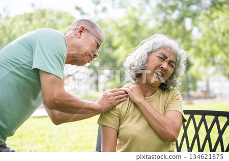 Old man holding or massaging pained or injury shoulder of woman sits on bench after exercise in park 134621875