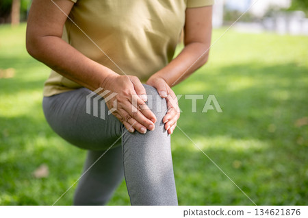 Old woman holding or massaging her pained or injury knee on bench after exercise or workout in park. 134621876