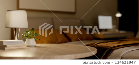Books with lamp and plant on round wooden table with blurred background of dim lit bedroom at night. Books with lamp and plant on round wooden table with blurred background of dim lit bedroom at night. 134621947
