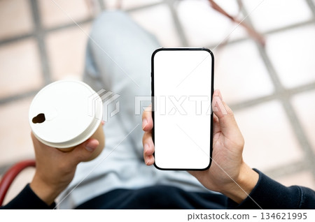 Hands holding coffee and white screen smartphone using application or social media as sits on bench. Hands holding coffee and white screen smartphone using application or social media as sits on bench. 134621995
