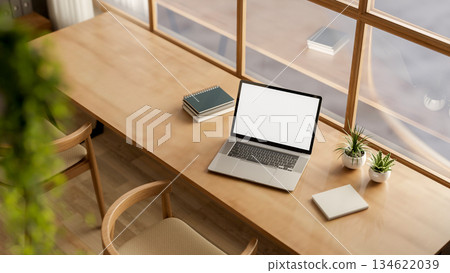 White screen laptop with book and plant on wooden table with chairs in a glass wall cafe or library. 134622039