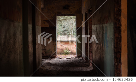 Sunlight enters through an open doorway in a long, narrow corridor. The worn walls show signs of age with peeling paint. Debris covers the floor, hinting at former use. Sunlight enters through an open doorway in a long, narrow corridor. The worn walls show signs of age with peeling paint. Debris covers the floor, hinting at former use. 134623426