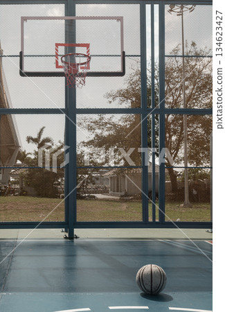 A worn basketball rests on the blue cement floor of an outdoor court with a basketball hoop, fiberglass backboard, and blue steel fence comprising the background. A worn basketball rests on the blue cement floor of an outdoor court with a basketball hoop, fiberglass backboard, and blue steel fence comprising the background. 134623427