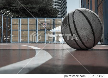 An old basketball lies on the cement in the foreground of a half-outdoor basketball court that features a basketball hoop, fiberglass backboard, and a blue steel fence. An old basketball lies on the cement in the foreground of a half-outdoor basketball court that features a basketball hoop, fiberglass backboard, and a blue steel fence. 134623429