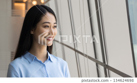 Chinese businesswoman standing near glass window, looks outside with smile Chinese businesswoman standing near glass window, looks outside with smile 134624698