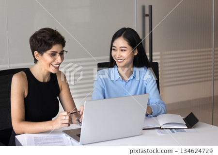 Two attractive multiethnic women colleagues sitting at desk with laptop 134624729
