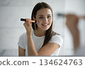 Young lady reflected in mirror using brush for facial visage 134624763