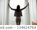 Beautiful female opening curtains on glass window wall admiring panorama 134624764