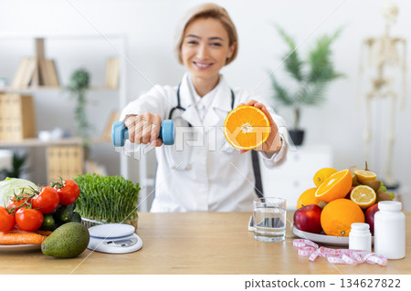 A smiling doctor holds a dumbbell and a sliced orange, promoting a healthy lifestyle with fruits and exercise. 134627822