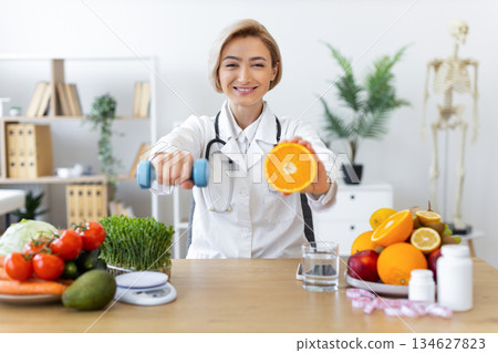 A smiling nutritionist holds an orange and a dumbbell, promoting healthy eating and exercise for a balanced lifestyle. 134627823