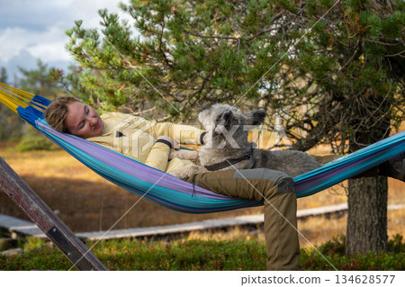 Woman relaxing in hammock with dog wearing outdoor clothing in forest nature 134628577