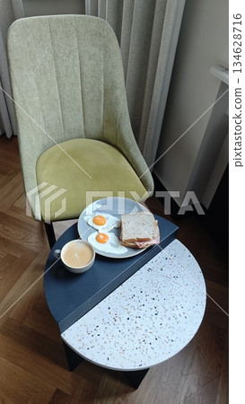 Plate with fried egg ham cheese sandwich and coffee on a small table in a cozy cafe setting. The image represents classic breakfast routine comfort food and relaxed morning lifestyle 134628716