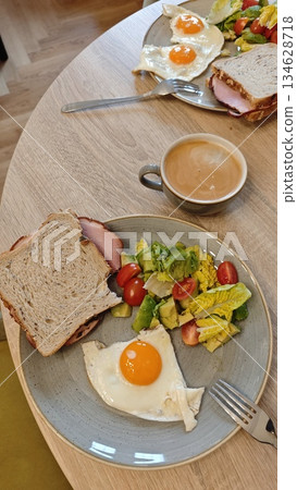 Plate with fried egg sandwich fresh salad and coffee served on a wooden table in a cozy cafe interior. The image represents balanced breakfast routine relaxed dining and healthy morning lifestyle 134628718