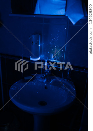 Ultraviolet black light reveals glowing traces of dirt, bacteria and contamination on a dark bathroom sink and soap dispenser, emphasizing hygiene and thorough cleaning. Ultraviolet black light reveals glowing traces of dirt, bacteria and contamination on a dark bathroom sink and soap dispenser, emphasizing hygiene and thorough cleaning. 134629800