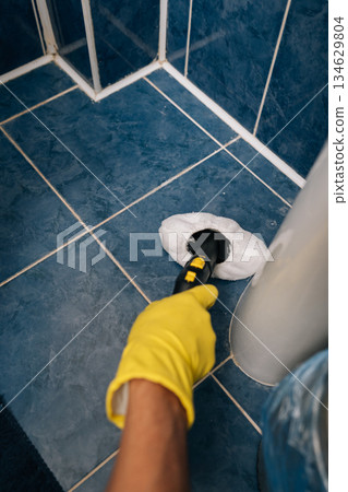 Hand in yellow protective glove using steam cleaner, deeply sanitizing blue floor tiles and white grout around base of toilet, ensuring thorough bathroom cleanliness. Concept of hygiene, household. Hand in yellow protective glove using steam cleaner, deeply sanitizing blue floor tiles and white grout around base of toilet, ensuring thorough bathroom cleanliness. Concept of hygiene, household. 134629804