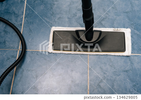 Top view of steam mop sanitizing blue ceramic bathroom tiles near a toilet, using hot vapor to remove dirt, grime and germs for a fresh, sparkling, hygienic floor surface. Concept of household chores. Top view of steam mop sanitizing blue ceramic bathroom tiles near a toilet, using hot vapor to remove dirt, grime and germs for a fresh, sparkling, hygienic floor surface. Concept of household chores. 134629805