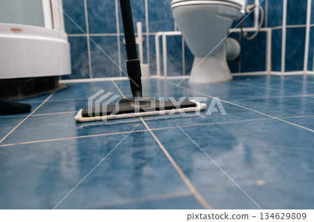 Close-up of steam mop sanitizing blue ceramic bathroom tiles near a toilet, using hot vapor to remove dirt, grime and germs for a fresh, sparkling, hygienic floor surface. Close-up of steam mop sanitizing blue ceramic bathroom tiles near a toilet, using hot vapor to remove dirt, grime and germs for a fresh, sparkling, hygienic floor surface. 134629809