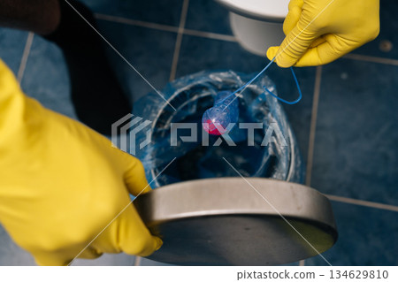 Top view of human hands in yellow protective gloves dropping used toilet bowl cleaner block into lined rubbish bin, highlighting bathroom hygiene, waste disposal and household cleaning routine. Top view of human hands in yellow protective gloves dropping used toilet bowl cleaner block into lined rubbish bin, highlighting bathroom hygiene, waste disposal and household cleaning routine. 134629810
