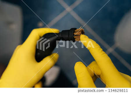 Close-up hands in yellow protective gloves holding checking steam cleaner with brush attachment, preparing to clean bathroom tiles for hygiene, sanitation and household chores. Close-up hands in yellow protective gloves holding checking steam cleaner with brush attachment, preparing to clean bathroom tiles for hygiene, sanitation and household chores. 134629811