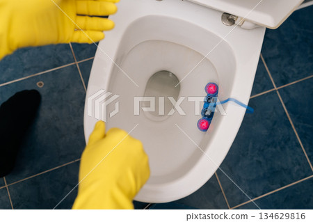 Top view of hands in yellow protective gloves placing toilet freshener block in clean bathroom, highlighting domestic hygiene, daily routine, household cleaning. Concept of hygiene and cleanliness. Top view of hands in yellow protective gloves placing toilet freshener block in clean bathroom, highlighting domestic hygiene, daily routine, household cleaning. Concept of hygiene and cleanliness. 134629816