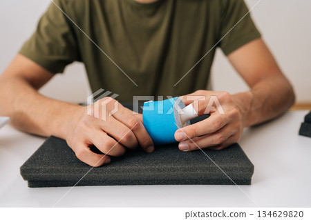 Male hand holding roll of kinesiology tape on table, preparing to apply for muscle, joint or tendon pain relief, representing self-care solution for aches and injuries. 134629820