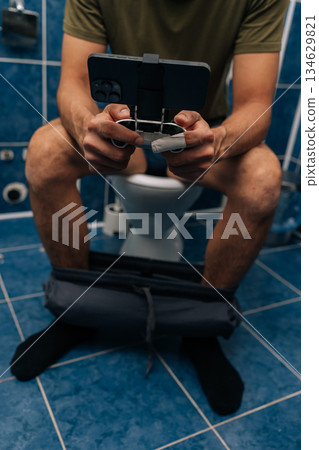Vertical cropped shot of young man holding smartphone with gamepad attachment and playing video games sitting on toilet, illustrating mobile gaming addiction, multitasking, privacy in bathroom. Vertical cropped shot of young man holding smartphone with gamepad attachment and playing video games sitting on toilet, illustrating mobile gaming addiction, multitasking, privacy in bathroom. 134629821
