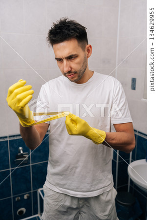 Man frustrated by torn rubber glove, experiencing cleaning problems and household chore difficulties in bathroom, highlighting disappointment with broken equipment and unexpected trouble. Man frustrated by torn rubber glove, experiencing cleaning problems and household chore difficulties in bathroom, highlighting disappointment with broken equipment and unexpected trouble. 134629955