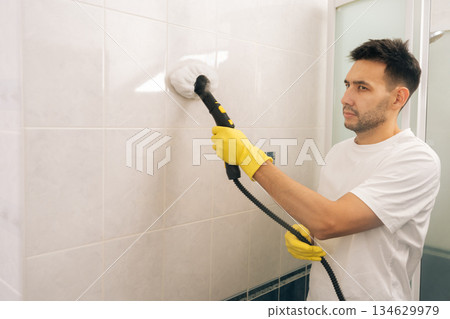Focused young man in protective yellow gloves cleaning bathroom wall tiles with powerful steam cleaner, ensuring deep hygiene, disinfection and sanitation in the domestic household environment. Focused young man in protective yellow gloves cleaning bathroom wall tiles with powerful steam cleaner, ensuring deep hygiene, disinfection and sanitation in the domestic household environment. 134629979