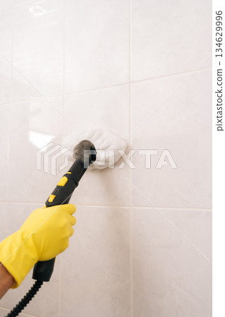 Close-up vertical shot of homemaker in yellow gloves using steam cleaner to deep-clean and sanitize white bathroom tiles, removing dirt and germs for a hygienic, comfortable home environment. Close-up vertical shot of homemaker in yellow gloves using steam cleaner to deep-clean and sanitize white bathroom tiles, removing dirt and germs for a hygienic, comfortable home environment. 134629996