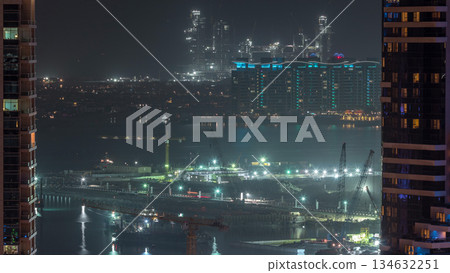 Aerial view to Dubai marina skyscrapers with construction site and Palm Jumeirah Island on background night timelapse. Aerial view to Dubai marina skyscrapers with construction site and Palm Jumeirah Island on background night timelapse. 134632251