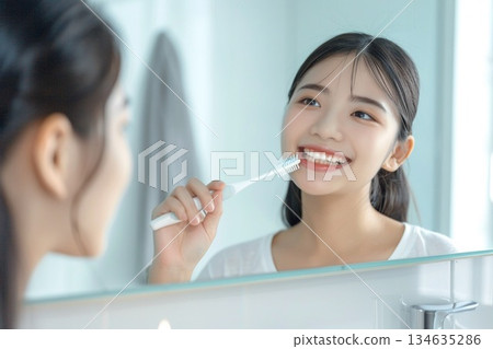 一位年輕女子在明亮的浴室裡拿著牙刷，露出潔白的牙齒，笑容燦爛。 134635286