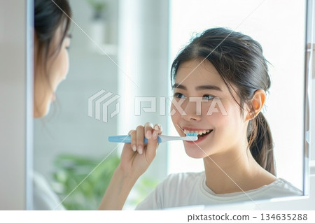 一位年輕女子在明亮的浴室裡拿著牙刷，露出潔白的牙齒，笑容燦爛。 134635288