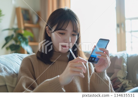 一位女士在家中客廳裡，一邊用智慧型手機接受線上醫療服務，一邊手裡拿著體溫計。 134635531