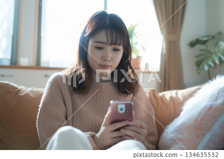 一位女士在家中客廳裡，一邊用智慧型手機接受線上醫療服務，一邊手裡拿著體溫計。 134635532