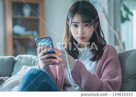 一位女士在家中客廳裡,一邊用智慧型手機接受線上醫療服務,一邊手裡拿著體溫計。 一位女士在家中客廳裡,一邊用智慧型手機接受線上醫療服務,一邊手裡拿著體溫計。 134635533