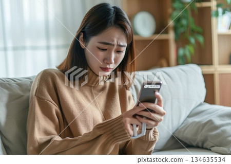 一位女士在家中客廳裡，一邊用智慧型手機接受線上醫療服務，一邊手裡拿著體溫計。 134635534