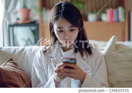 一位女士在家中客廳裡,一邊用智慧型手機接受線上醫療服務,一邊手裡拿著體溫計。 一位女士在家中客廳裡,一邊用智慧型手機接受線上醫療服務,一邊手裡拿著體溫計。 134635535