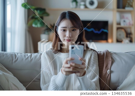 一位女士在家中客廳裡,一邊用智慧型手機接受線上醫療服務,一邊手裡拿著體溫計。 一位女士在家中客廳裡,一邊用智慧型手機接受線上醫療服務,一邊手裡拿著體溫計。 134635536