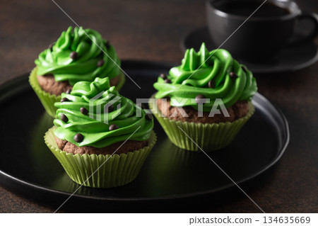 Three chocolate cupcakes with green whipped cream and chocolate sprinkles on dark brown background. St. Patrick's Day. Close up. Three chocolate cupcakes with green whipped cream and chocolate sprinkles on dark brown background. St. Patrick's Day. Close up. 134635669