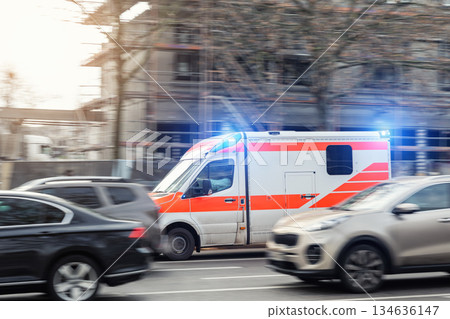 Ambulance flash emergency lights driving fast busy city traffic jam motion blur among cars urban road. Urgent medical response scene shows rescue transport public safety service rapid European city 134636147