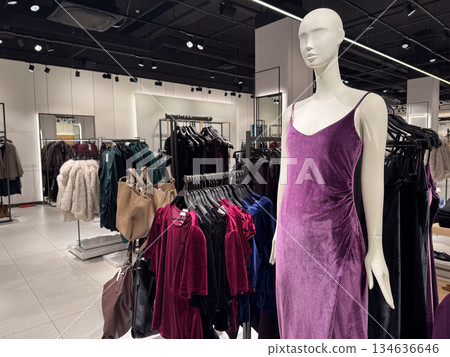 A modern fashion store interior features a mannequin dressed in a purple velvet dress surrounded by clothing racks A modern fashion store interior features a mannequin dressed in a purple velvet dress surrounded by clothing racks 134636646