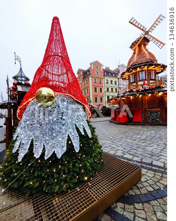 Large decorative gnome figure, with a red, cone-shaped hat, silver beard, and a spherical gold nose, crafted from evergreen branches with lights and wooden Christmas stall topped with windmill at 134636916
