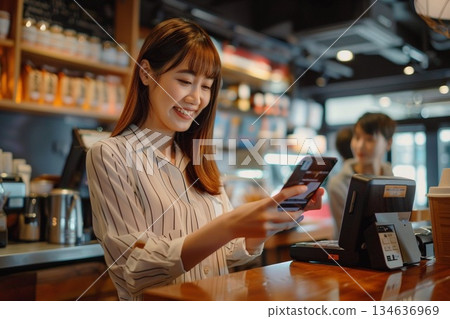 一位微笑的女士在咖啡館櫃檯使用智慧型手機進行無現金支付。 134636969