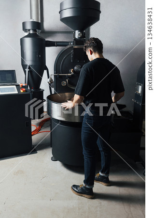 Full length of a male worker at an industrial roasting coffee machine. Young man controlling the roasting coffee process. Full length of a male worker at an industrial roasting coffee machine. Young man controlling the roasting coffee process. 134638431