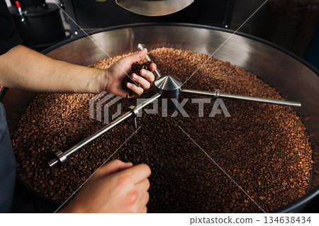 Unrecognizable male grabs roasted coffee beans from the machine drum. High detail shot of a male hand holding fresh roasted beans. Unrecognizable male grabs roasted coffee beans from the machine drum. High detail shot of a male hand holding fresh roasted beans. 134638434