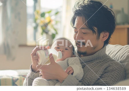 A kind father holding his baby and feeding him milk from a bottle in a bright living room 134638589
