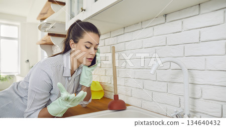 Young girl talking by the phone advising how to unclog a sink drain in the kitchen at home. 134640432