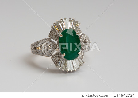 A gold ring featuring an oval cut green emerald surrounded by baguette cut diamonds on a clean white background. A gold ring featuring an oval cut green emerald surrounded by baguette cut diamonds on a clean white background. 134642724