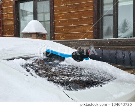 Woman clearing snow from her car windshield during winter. Preparing vehicle for travel in cold weather. Winter season car maintenance. Woman clearing snow from her car windshield during winter. Preparing vehicle for travel in cold weather. Winter season car maintenance. 134643720