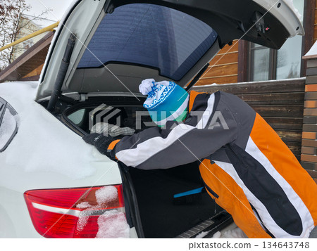 Adult man in winter clothing with knitted hat puts something into white car trunk in snowy day. Winter season concept. Adult man in winter clothing with knitted hat puts something into white car trunk in snowy day. Winter season concept. 134643748